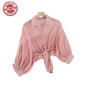 Peut inclure: Un chemisier rose clair et transparent avec de longues manches volumineuses et une fermeture à nouer. Le chemisier a un col et une fermeture boutonnée. Les manches ont des poignets ajustés. Le tissu semble léger et fluide.
