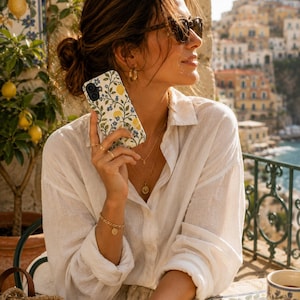 Pode incluir: Uma capa de telefone branca com um padr&atilde;o floral e de lim&atilde;o &eacute; segurada por uma pessoa. A capa apresenta lim&otilde;es amarelos, flores azuis e folhas verdes sobre um fundo creme. A pessoa est&aacute; usando &oacute;culos de sol e uma camisa branca.