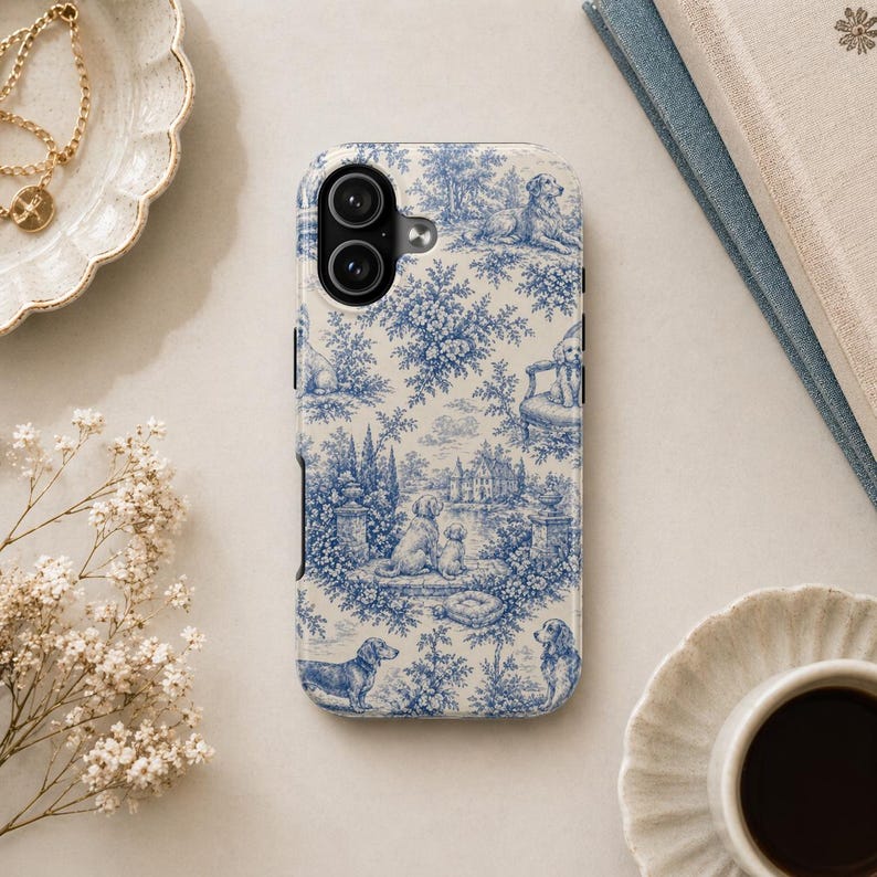K&ouml;nnte beinhalten: Eine Handyh&uuml;lle mit blau-wei&szlig;em Toile-de-Jouy-Muster mit Hunden, B&auml;umen und Geb&auml;uden. Die H&uuml;lle hat einen Kameraausschnitt und einen schwarzen Rand. Das Design erinnert an klassische franz&ouml;sische Landszenen.