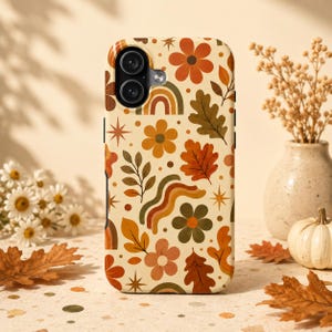 May include: A phone case featuring a cream background with a repeating pattern of orange, brown, and olive green flowers, oak leaves, and rainbow arches. The design includes small starburst shapes and dots scattered throughout the surface.