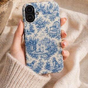 K&ouml;nnte beinhalten: Eine Handyh&uuml;lle mit blau-wei&szlig;em Toile-de-Jouy-Muster mit Hunden in einer Gartenlandschaft. Die H&uuml;lle hat einen schwarzen Rand und einen Ausschnitt f&uuml;r eine Dual-Kamera. Das Design umfasst B&auml;ume, Geb&auml;ude und verschiedene Hunderassen.