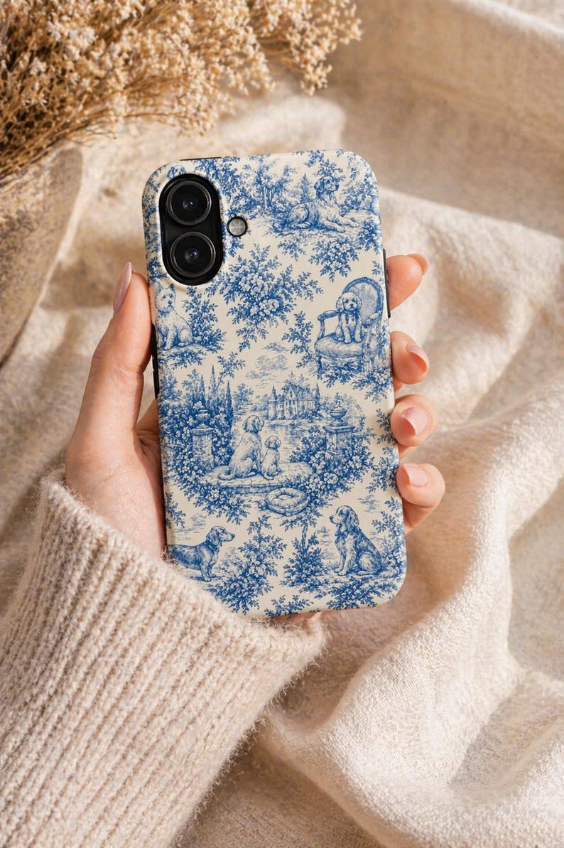 K&ouml;nnte beinhalten: Eine Handyh&uuml;lle mit blau-wei&szlig;em Toile-de-Jouy-Muster mit Hunden in einer Gartenlandschaft. Die H&uuml;lle hat einen schwarzen Rand und einen Ausschnitt f&uuml;r eine Dual-Kamera. Das Design umfasst B&auml;ume, Geb&auml;ude und verschiedene Hunderassen.