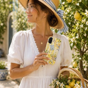 May include: A phone case featuring a yellow and white striped pattern with illustrations of lemons, a straw hat, a basket, and a book. The case has a dual-camera cutout at the top left and a glossy finish, displaying a vibrant citrus-themed design.