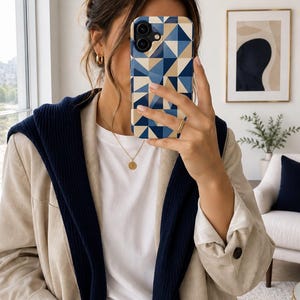May include: A smartphone case featuring a geometric design of blue and beige triangles. The case is held up in front of a person's face. The person is wearing a white t-shirt, a beige blazer, and a navy blue sweater draped over the shoulders.