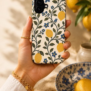 Pode incluir: Uma capa de telefone com fundo creme com um padr&atilde;o de lim&otilde;es amarelos, flores azuis e folhas verdes. A capa &eacute; segurada em uma m&atilde;o, com um anel e uma pulseira dourados vis&iacute;veis. Uma tigela com desenhos florais azuis e um lim&atilde;o no fundo.