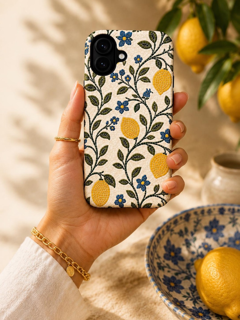 Pode incluir: Uma capa de telefone com fundo creme com um padr&atilde;o de lim&otilde;es amarelos, flores azuis e folhas verdes. A capa &eacute; segurada em uma m&atilde;o, com um anel e uma pulseira dourados vis&iacute;veis. Uma tigela com desenhos florais azuis e um lim&atilde;o no fundo.
