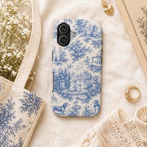 K&ouml;nnte beinhalten: Eine Handyh&uuml;lle mit blau-wei&szlig;em Toile-de-Jouy-Design mit Hunden, B&auml;umen und Geb&auml;uden. Die H&uuml;lle ist umgeben von einer goldenen Halskette mit Herzanh&auml;nger, einem goldenen Ring und einer Tragetasche mit passendem Muster.
