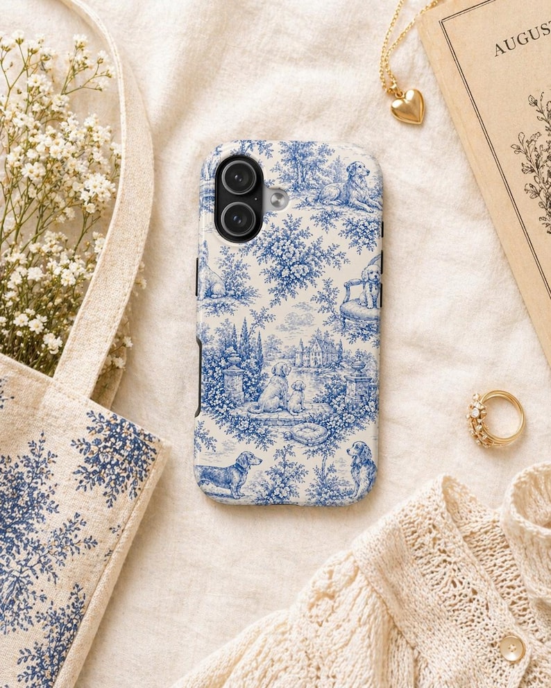 K&ouml;nnte beinhalten: Eine Handyh&uuml;lle mit blau-wei&szlig;em Toile-de-Jouy-Design mit Hunden, B&auml;umen und Geb&auml;uden. Die H&uuml;lle ist umgeben von einer goldenen Halskette mit Herzanh&auml;nger, einem goldenen Ring und einer Tragetasche mit passendem Muster.