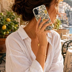 Pode incluir: Um smartphone com uma capa de design floral com lim&otilde;es amarelos e flores azuis. O telefone &eacute; segurado perto da orelha. A pessoa est&aacute; vestindo uma camisa branca abotoada e joias de ouro. O fundo mostra uma cena costeira com lim&otilde;es.