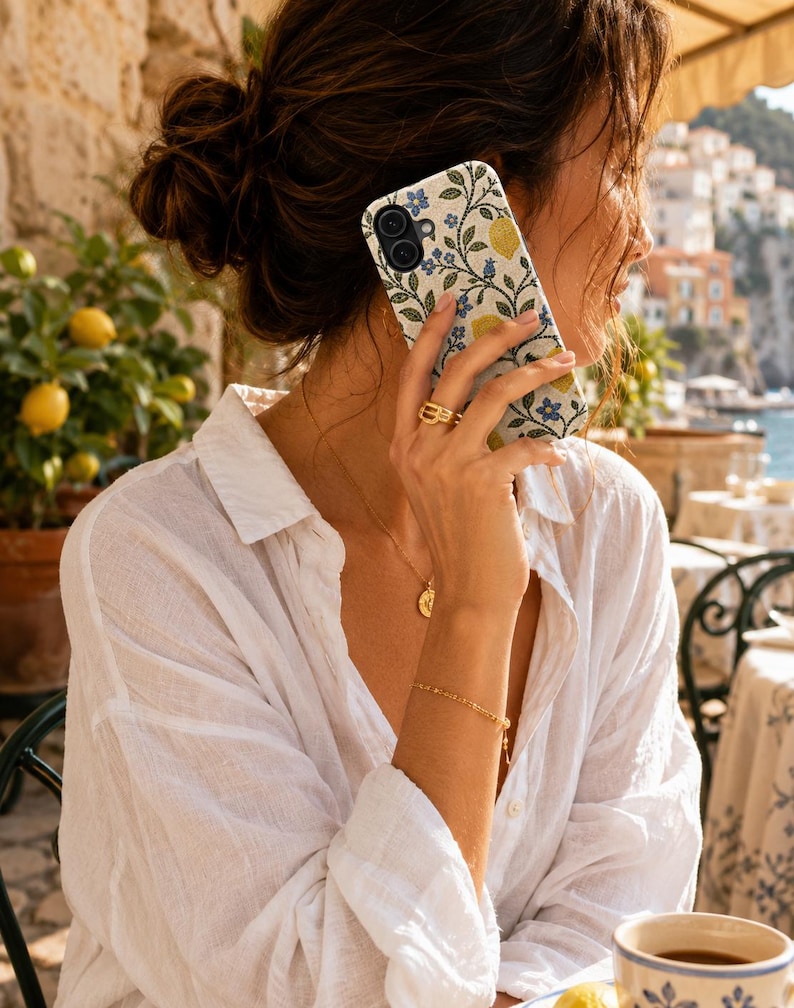 Pode incluir: Um smartphone com uma capa de design floral com lim&otilde;es amarelos e flores azuis. O telefone &eacute; segurado perto da orelha. A pessoa est&aacute; vestindo uma camisa branca abotoada e joias de ouro. O fundo mostra uma cena costeira com lim&otilde;es.