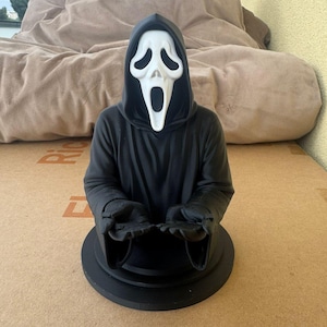 Könnte beinhalten: Eine schwarze Büste der Ghostface-Maske aus der Horrorfilmreihe Scream. Die Maske ist weiß mit schwarzen Augen- und Mundöffnungen, vor einem schwarzen Kapuzenmantel. Die Büste steht auf einem schwarzen kreisförmigen Sockel.