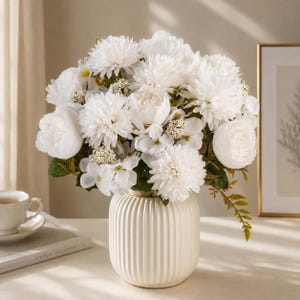 May include: A bouquet of white artificial flowers, including chrysanthemums, peonies, and small filler blossoms with green leaves, arranged in a round, cream-colored ceramic vase with vertical ribbed textures and a wide, flat base.