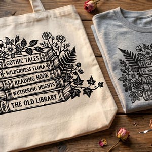 May include: A light-colored tote bag and a gray t-shirt, both featuring a black floral design and stacked books with titles like "Gothic Tales" and "Wuthering Heights." The items are displayed on a wooden surface.