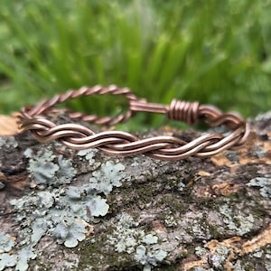 May include: A copper-colored wire bracelet with a braided design. The bracelet is resting on a piece of wood with lichen and moss. The background is blurred green grass.