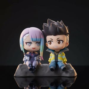 Puede incluir: Dos figuras de estilo chibi de personajes de Cyberpunk 2077. La figura femenina tiene cabello lavanda con reflejos azules y verdes, vistiendo un atuendo morado. La figura masculina tiene cabello negro y una chaqueta amarilla. Están sentados en una roca gris.