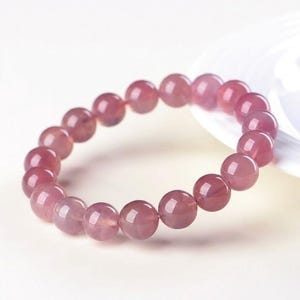 May include: A pink gemstone bracelet featuring round beads. The beads have a translucent appearance and a soft, rosy hue. The bracelet is displayed on a white surface, showcasing its color and design. The bracelet is a piece of jewelry.