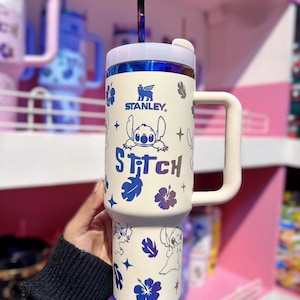 May include: Cream-colored Stanley tumbler featuring Stitch from Lilo & Stitch. The tumbler has a white handle and lid, with a blue and purple straw. The design includes Stitch illustrations, blue flowers, and the word "Stitch".