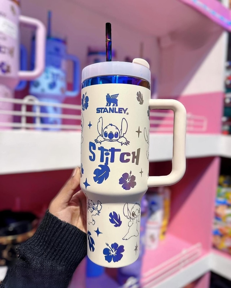 May include: Cream-colored Stanley tumbler featuring Stitch from Lilo & Stitch. The tumbler has a white handle and lid, with a blue and purple straw. The design includes Stitch illustrations, blue flowers, and the word "Stitch".
