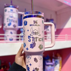 May include: A pink Stanley tumbler featuring Stitch character designs and the word "Stitch". The tumbler has a handle, a clear lid, and a silver straw. The Stanley logo is visible. The design includes blue and iridescent accents.