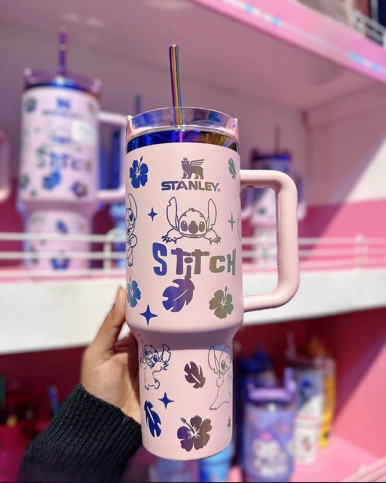 May include: A pink Stanley tumbler featuring Stitch character designs and the word "Stitch". The tumbler has a handle, a clear lid, and a silver straw. The Stanley logo is visible. The design includes blue and iridescent accents.