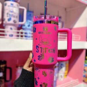 May include: A bright pink Stanley tumbler with a handle, featuring a Stitch design. The tumbler has a clear lid and a rainbow-colored straw. The design includes Stitch, flowers, and stars. The word "Stitch" is printed on the tumbler.