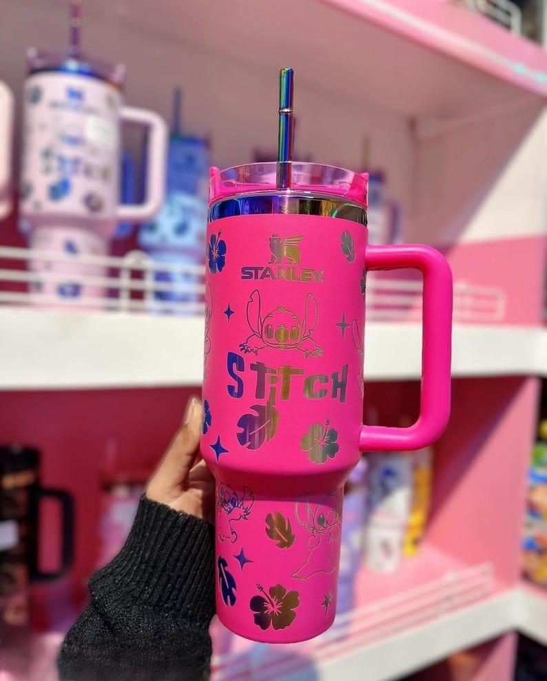 May include: A bright pink Stanley tumbler with a handle, featuring a Stitch design. The tumbler has a clear lid and a rainbow-colored straw. The design includes Stitch, flowers, and stars. The word "Stitch" is printed on the tumbler.