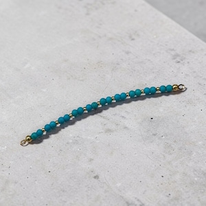 May include: A delicate strand of turquoise beads with gold-colored accents. The beads are strung together, forming a curved line. The ends of the strand have small gold-colored loops. The piece is set against a neutral, light gray background.