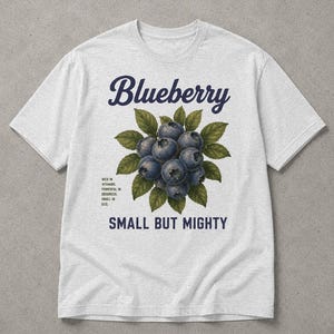 Op de afbeelding: Een lichtgrijs t-shirt met een centrale afbeelding van een tros bosbessen met groene bladeren. De tekst luidt bovenaan Blueberry, onderaan Small But Mighty en links Rich in vitamins, powerful in goodness, small in size.