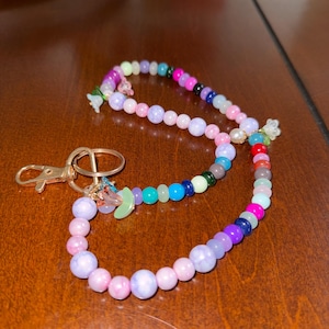May include: A beaded keychain with a gold clasp. The beads are in various colours, including pink, purple, blue, green, and white. Small flower and moon charms are attached to the keychain.