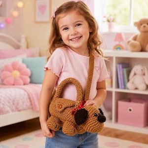 Può includere: Una borsa a forma di cane all'uncinetto marrone con un fiocco rosso e dettagli neri. La borsa ha un manico e una tracolla. La ragazza indossa una maglietta rosa e jeans blu. Lo sfondo include un letto, scaffali e giocattoli.