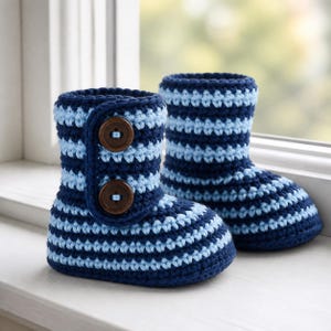 Op de afbeelding: Een paar handgehaakte slofjes met horizontale strepen in donker marineblauw en lichtblauw garen. De voorkant van de slofjes heeft een verticale flap die is vastgezet met twee ronde, donkerbruine houten knopen met zichtbare stiksels.