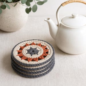 May include: A stack of circular crochet coasters featuring a central dark gray star, surrounded by a ring of rust-colored petals and an outer border of cream yarn. The sides of the coasters are constructed with alternating rows of dark gray and cream yarn.