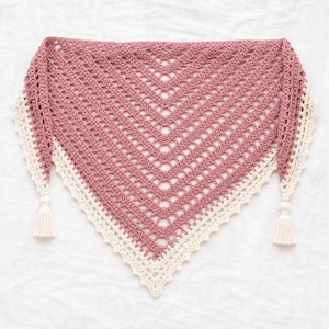 Pode incluir: Um xale triangular de crochê com um centro rosa antigo e um padrão de pontos abertos. As bordas são finalizadas com um acabamento festonado na cor creme e uma borla creme correspondente presa a cada um dos dois cantos laterais curtos.