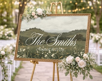 May include: A rectangular wedding sign in a gold ornate frame featuring a landscape painting of green hills and a meadow. The text reads The Smiths and August 8, 2026 in white script. The sign is decorated with white roses and greenery on the corners.