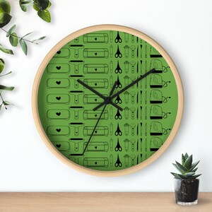 Puede incluir: Un reloj de pared redondo con marco de madera clara. La esfera verde del reloj presenta un patrón repetido de herramientas de artesanía negras, incluyendo tijeras, máquinas de corte y prensas térmicas. Las manecillas son negras.