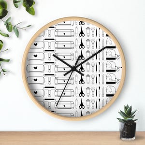 Puede incluir: Reloj de pared redondo con marco de madera clara. La esfera del reloj presenta un patrón repetido de herramientas de artesanía en blanco y negro, incluyendo tijeras, máquinas de corte y prensas térmicas. Las manecillas del reloj son negras.