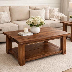 May include: A rectangular, dark brown wooden coffee table with a lower shelf. The table is in a living room setting, with a beige sofa and a white vase of flowers on top. A candle and a book are also on the table.