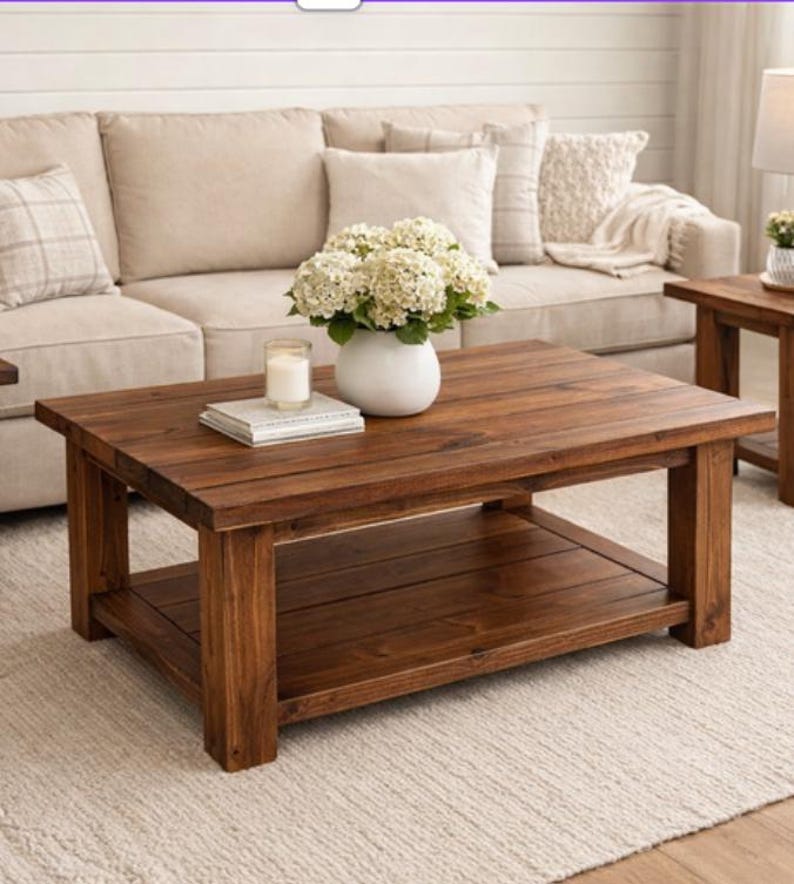 May include: A rectangular, dark brown wooden coffee table with a lower shelf. The table is in a living room setting, with a beige sofa and a white vase of flowers on top. A candle and a book are also on the table.