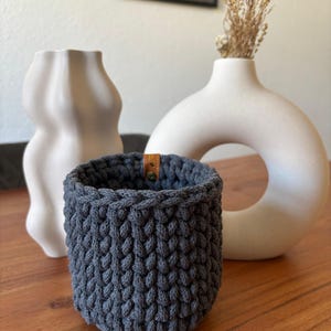Peut inclure: Un pot en crochet gris foncé avec une poignée en cuir marron. Le pot est posé sur une table en bois avec deux vases en céramique blanche.