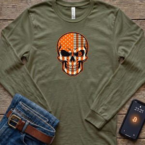 May include: An olive green long-sleeve shirt featuring a graphic of a human skull. The skull is split into two patterns: one side displays white stars on an orange background, and the other side features orange binary code on a white background.