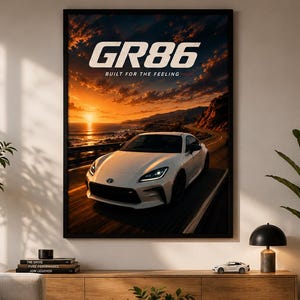 May include: A framed wall poster featuring a white sports car driving along a coastal road at sunset. The text GR86 and BUILT FOR THE FEELING is printed in white at the top. The scene depicts a vibrant orange sky over the ocean and mountains.