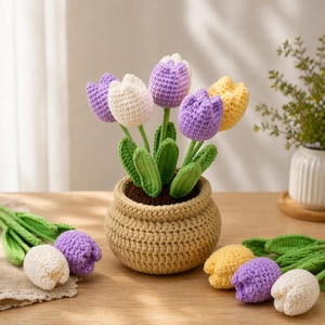 May include: A crocheted flower arrangement featuring tulips in a woven pot. The tulips are in shades of purple, white, and yellow, with green stems and leaves. The pot is a light brown color, and the arrangement is set on a wooden surface.