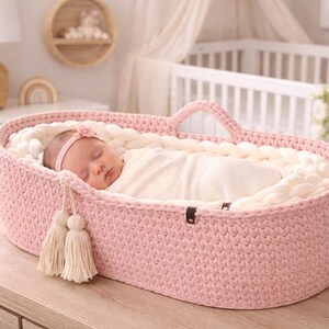 May include: A sleeping baby in a pink crocheted bassinet with a white interior. The bassinet has a handle and two decorative tassels. The baby is wrapped in a white blanket and wearing a pink headband. The bassinet is on a wooden surface.