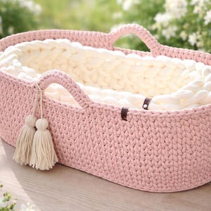 May include: A pink, oval-shaped crochet baby basket with two handles. The interior is lined with a thick, cream-colored yarn. Two cream-colored tassels hang from one handle. The basket is approximately 20 inches long and 12 inches wide.