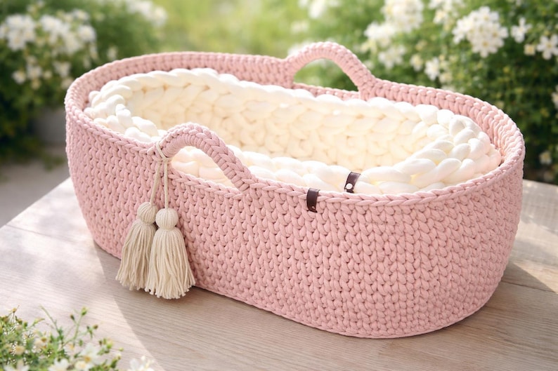 May include: A pink, oval-shaped crochet baby basket with two handles. The interior is lined with a thick, cream-colored yarn. Two cream-colored tassels hang from one handle. The basket is approximately 20 inches long and 12 inches wide.