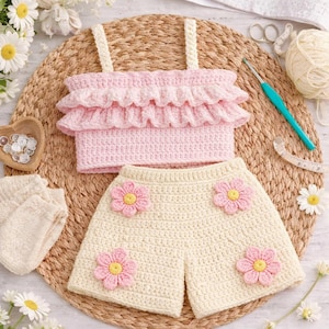 May include: A handmade crochet outfit for a child, featuring a pink ruffled top with cream straps and matching cream shorts adorned with pink and yellow flower appliqués. The set is displayed on a woven mat with crochet supplies.