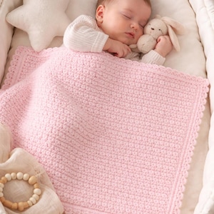 Puede incluir: Un bebé dormido está acurrucado en una cuna blanca, cubierto con una suave manta rosa de ganchillo. Una almohada blanca en forma de estrella y un conejito de peluche también están presentes. Un mordedor de madera descansa sobre una tela color crema.