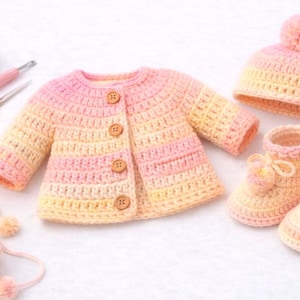 May include: A hand-crocheted baby set in pink and yellow hues. The set comprises a button-up jacket, a beanie with a pom-pom, and booties with bows. Yarn, a crochet hook, and scissors are also visible, all in a soft colour palette.