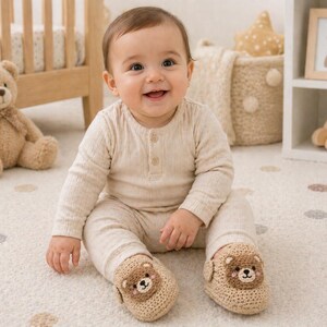 Puede incluir: Un beb&eacute; sonriente viste una camisa beige de manga larga acanalada y pantalones a juego. El beb&eacute; lleva botines de oso de ganchillo beige. Un osito de peluche y una cuna est&aacute;n en el fondo.
