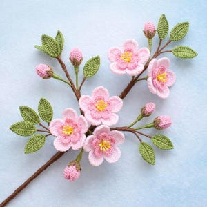 May include: A crocheted branch featuring pink cherry blossoms with yellow centres, green leaves, and pink buds. The handmade floral design is set against a light blue background. The brown stem adds a natural touch to the delicate craft.